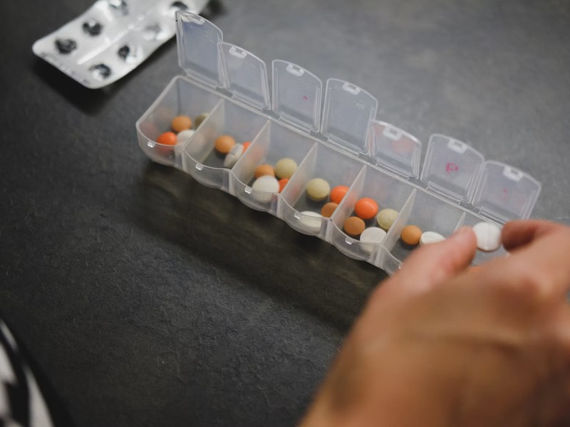 A hand sorting tablets into a weekly pill organizer beside a blister pack
